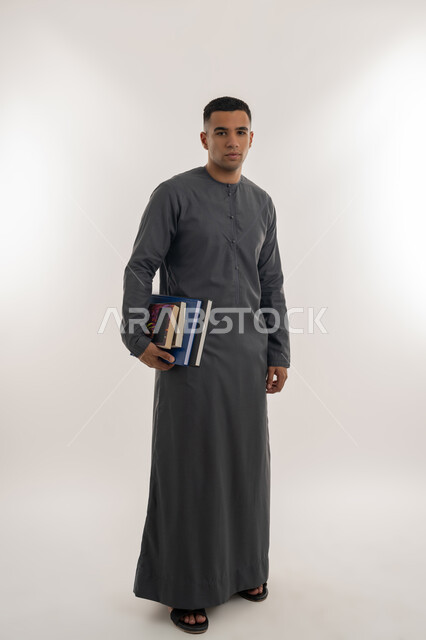 المناهج والمواد التعليمية, الاستعداد للعام الجامعي الجديد, التعليم العالي في الجامعات في الإمارات العربية المتحدة, بورتريه لطالب جامعي عربي خليجي إماراتي يرتدي الكندورة ينظر إلى الكاميرا ويحمل بيده الكتب الجامعية, الجسم بالطول الكامل, خلفية رمادية
