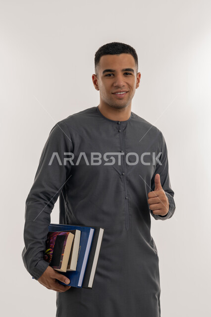 تعابير الجودة و الامتياز, التعليم الجامعي في الإمارات العربية المتحدة, النظر إلى الكاميرا بإيماءات السعادة والسرور, بورتريه لطالب جامعي عربي خليجي إماراتي يرتدي الكندورة يحمل بيده الكتب الجامعية ويشير بإصبع الإبهام للأعلى, خلفية رمادية