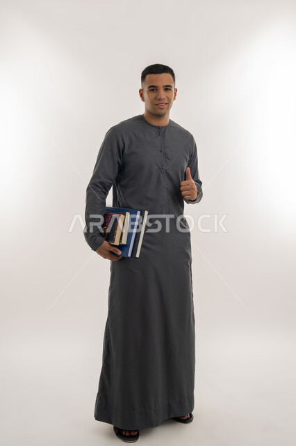 تعابير الجودة و الامتياز, التعليم الجامعي في الإمارات العربية المتحدة, النظر إلى الكاميرا بإيماءات السعادة والسرور, بورتريه لطالب جامعي عربي خليجي إماراتي يرتدي الكندورة يحمل بيده الكتب الجامعية ويشير بإصبع الإبهام للأعلى, الجسم بالطول الكامل, خلفية رمادية