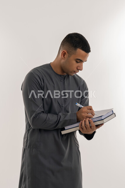 التعليم العالي في الجامعات والكليات الأكاديمية في الإمارات العربية المتحدة, دراسة المحاضرات والمقررات, مفهوم الاجتهاد والتفوق, بورتريه لطالب جامعي عربي خليجي إماراتي يرتدي الكندورة يحمل بيده الكتب الجامعية ويقوم بالكتابة ومراجعة الدروس اليومية, خلفية رمادية