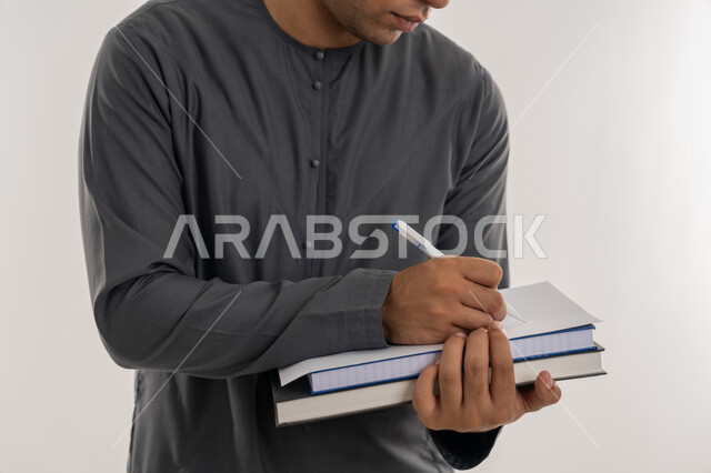 دراسة المحاضرات والمقررات, التعليم العالي في الجامعات والكليات الأكاديمية في الإمارات العربية المتحدة, مفهوم الاجتهاد والتفوق, بورتريه مقرب لطالب جامعي عربي خليجي إماراتي يرتدي الكندورة يحمل بيده الكتب ويقوم بالكتابة ومراجعة الدروس اليومية, خلفية رمادية