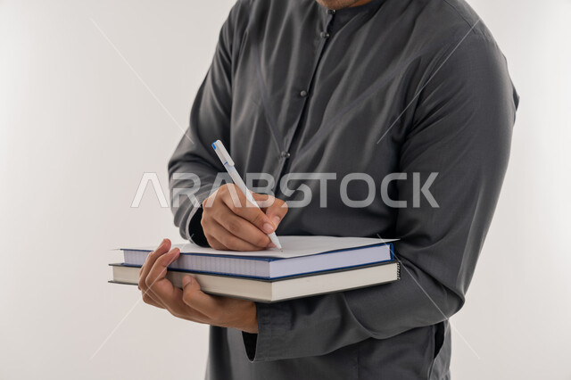 دراسة المحاضرات والمقررات, التعليم العالي في الجامعات والكليات الأكاديمية في الإمارات العربية المتحدة, مفهوم الاجتهاد والتفوق, بورتريه مقرب لطالب جامعي عربي خليجي إماراتي يرتدي الكندورة يحمل بيده الكتب ويقوم بالكتابة ومراجعة الدروس اليومية, خلفية رمادية