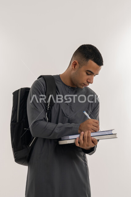 التعليم العالي في الجامعات والكليات الأكاديمية في الإمارات العربية المتحدة, دراسة المحاضرات والمقررات, مفهوم الاجتهاد والتفوق, بورتريه لطالب جامعي عربي خليجي إماراتي يرتدي الكندورة يحمل بيده الكتب الجامعية ويقوم بالكتابة ومراجعة الدروس اليومية, خلفية رمادية