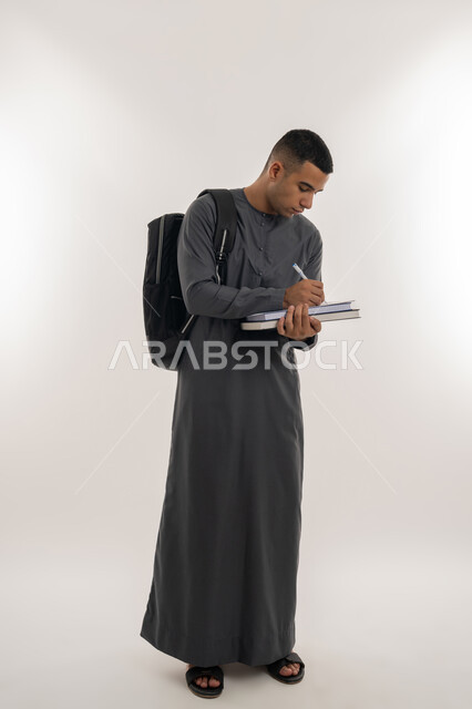 مراجعة الدروس اليومية, التعليم الجامعي في الإمارات العربية المتحدة, مفهوم الاجتهاد والتفوق, بورتريه لطالب جامعي عربي خليجي إماراتي يرتدي الكندورة يحمل حقيبة على ظهره وبيده الكتب الجامعية ويقوم بدراسة المحاضرات, الجسم بالطول الكامل, خلفية رمادية