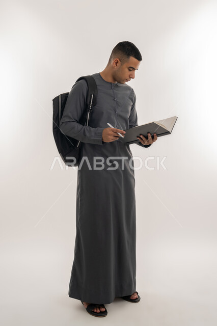 مفهوم الاجتهاد والتفوق, مراجعة الدروس اليومية, التعليم الجامعي في الإمارات العربية المتحدة, بورتريه لطالب جامعي عربي خليجي إماراتي يرتدي الكندورة يحمل حقيبة على ظهره وبيده كتاب ويقوم بدراسة المحاضرات, الجسم بالطول الكامل, خلفية رمادية