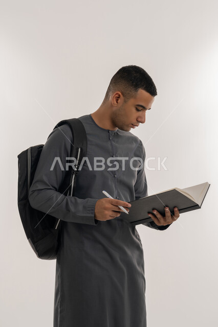 التعليم الجامعي في الإمارات العربية المتحدة, مراجعة الدروس اليومية, مفهوم الاجتهاد والتفوق, بورتريه لطالب جامعي عربي خليجي إماراتي يرتدي الكندورة يحمل حقيبة على ظهره وبيده كتاب ويقوم بدراسة المحاضرات, خلفية رمادية