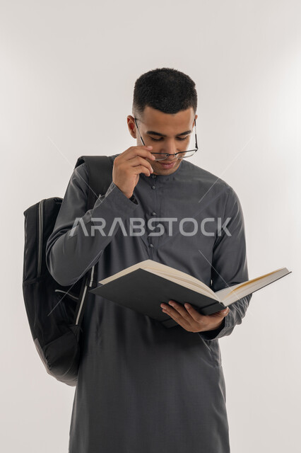 التعليم الجامعي في الإمارات العربية المتحدة, مراجعة الدروس اليومية, مفهوم الاجتهاد والتفوق, بورتريه لطالب جامعي عربي خليجي إماراتي يرتدي الكندورة يحمل حقيبة على ظهره وبيده كتاب ويقوم بدراسة المحاضرات, خلفية رمادية