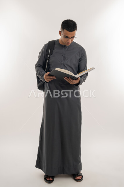 مفهوم الاجتهاد والتفوق, مراجعة الدروس اليومية, التعليم الجامعي في الإمارات العربية المتحدة, بورتريه لطالب جامعي عربي خليجي إماراتي يرتدي الكندورة يحمل حقيبة على ظهره وبيده كتاب ويقوم بدراسة المحاضرات, الجسم بالطول الكامل, خلفية رمادية