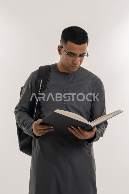 التعليم الجامعي في الإمارات العربية المتحدة, مراجعة الدروس اليومية, مفهوم الاجتهاد والتفوق, بورتريه لطالب جامعي عربي خليجي إماراتي يرتدي الكندورة يحمل حقيبة على ظهره وبيده كتاب ويقوم بدراسة المحاضرات, خلفية رمادية