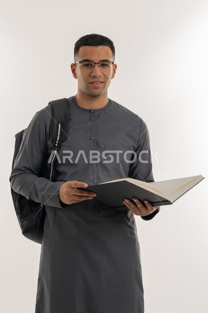 النظر إلى الكاميرا بإيماءات السعادة والسرور, مراجعة الدروس اليومية, التعليم الجامعي في الإمارات العربية المتحدة, مفهوم الاجتهاد والتفوق, بورتريه لطالب جامعي عربي خليجي إماراتي يرتدي الكندورة يحمل حقيبة على ظهره وبيده كتاب ويقوم بدراسة المحاضرات, خلفية رمادية