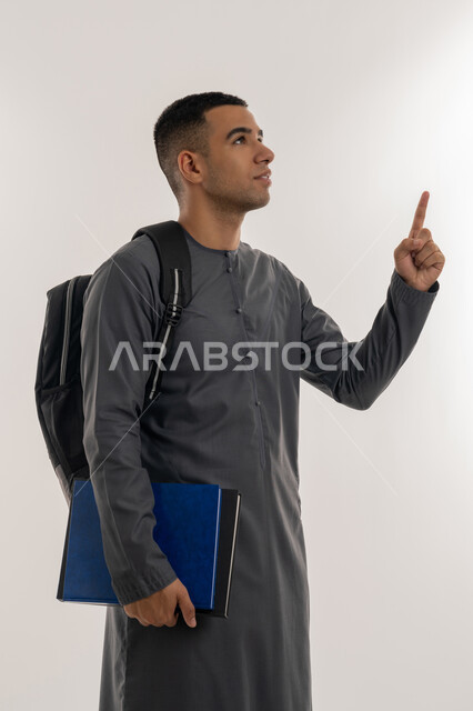 الاستعداد للعام الجامعي الجديد, الإشارة بإصبع السبابة إلى شيء ما, التعليم الاكاديمي في الإمارات العربية المتحدة, بورتريه لطالب جامعي عربي خليجي إماراتي يرتدي الكندورة ينظر إلى الأعلى ويحمل حقيبة على ظهره والكتب بيديه, خلفية رمادية