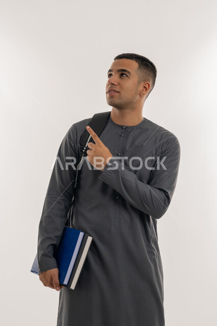 الإشارة بإصبع السبابة إلى شيء ما, الاستعداد للعام الجامعي الجديد, التعليم الاكاديمي في الإمارات العربية المتحدة, بورتريه لطالب جامعي عربي خليجي إماراتي يرتدي الكندورة ينظر إلى الأعلى ويحمل حقيبة على ظهره والكتب بيديه, خلفية رمادية