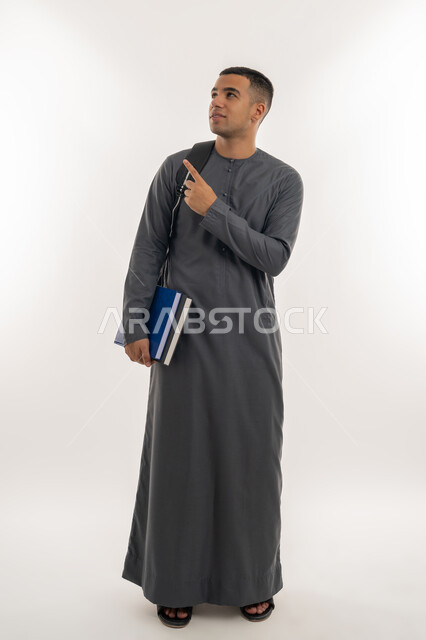 الإشارة بإصبع السبابة إلى شيء ما, الاستعداد للعام الجامعي الجديد, التعليم الاكاديمي في الإمارات العربية المتحدة, بورتريه لطالب جامعي عربي خليجي إماراتي يرتدي الكندورة ينظر إلى الأعلى ويحمل حقيبة على ظهره والكتب بيديه, الجسم بالطول الكامل, خلفية رمادية
