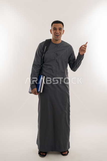 النظر إلى الكاميرا بإيماءات السعادة والسرور, الاستعداد للعام الجامعي الجديد, التعليم الاكاديمي في الإمارات العربية المتحدة, بورتريه لطالب جامعي عربي خليجي إماراتي يرتدي الكندورة يحمل بيده الكتب ويشير بإصبع السبابة للأعلى, الجسم بالطول الكامل, خلفية رمادية