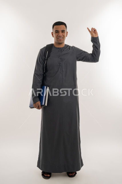 النظر إلى الكاميرا بإيماءات السعادة والسرور, الاستعداد للعام الجامعي الجديد, التعليم الاكاديمي في الإمارات العربية المتحدة, بورتريه لطالب جامعي عربي خليجي إماراتي يرتدي الكندورة يحمل بيده الكتب ويشير بإصبع السبابة للأعلى, الجسم بالطول الكامل, خلفية رمادية