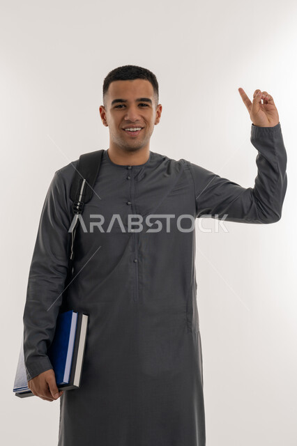 الاستعداد للعام الجامعي الجديد, التعليم الاكاديمي في الإمارات العربية المتحدة, الإشارة بإصبع السبابة إلى شيء ما, بورتريه لطالب جامعي عربي خليجي إماراتي يرتدي الكندورة ينظر إلى الكاميرا ويحمل حقيبة على ظهره والكتب بيديه, خلفية رمادية