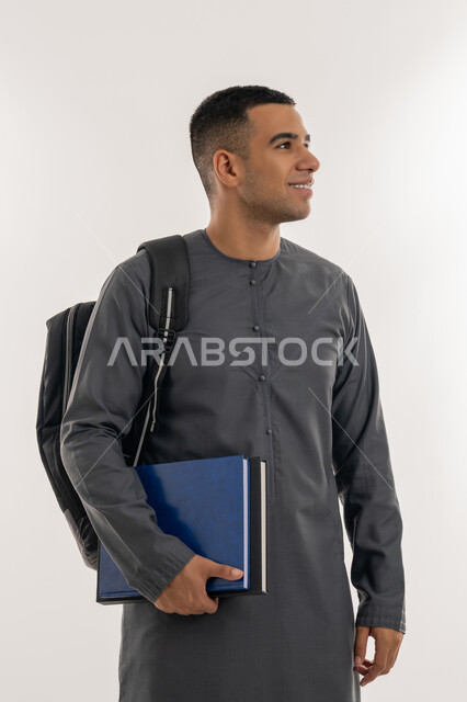 النظر بإيماءات السعادة والسرور, الاستعداد للعام الجامعي الجديد, التعليم العالي في الجامعات والكليات الأكاديمية الإماراتية, بورتريه لطالب جامعي عربي خليجي إماراتي يرتدي الكندورة يحمل حقيبة على ظهره وبيديه الكتب, خلفية رمادية