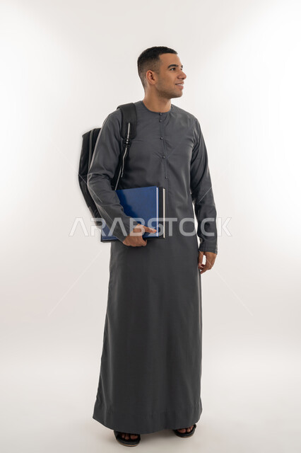 النظر بإيماءات السعادة والسرور, الاستعداد للعام الجامعي الجديد, التعليم العالي في الجامعات والكليات الأكاديمية الإماراتية, بورتريه لطالب جامعي عربي خليجي إماراتي يرتدي الكندورة يحمل حقيبة على ظهره وبيديه الكتب, الجسم بالطول الكامل, خلفية رمادية