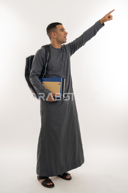 الإشارة بإصبع السبابة إلى شيء ما, الاستعداد للعام الجامعي الجديد, التعليم الاكاديمي في الإمارات العربية المتحدة, بورتريه لطالب جامعي عربي خليجي إماراتي يرتدي الكندورة ينظر إلى الأعلى ويحمل حقيبة على ظهره والكتب بيديه, الجسم بالطول الكامل, خلفية رمادية