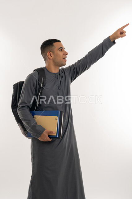 الإشارة بإصبع السبابة إلى شيء ما, الاستعداد للعام الجامعي الجديد, التعليم الاكاديمي في الإمارات العربية المتحدة, بورتريه لطالب جامعي عربي خليجي إماراتي يرتدي الكندورة ينظر إلى الأعلى ويحمل حقيبة على ظهره والكتب بيديه, خلفية رمادية