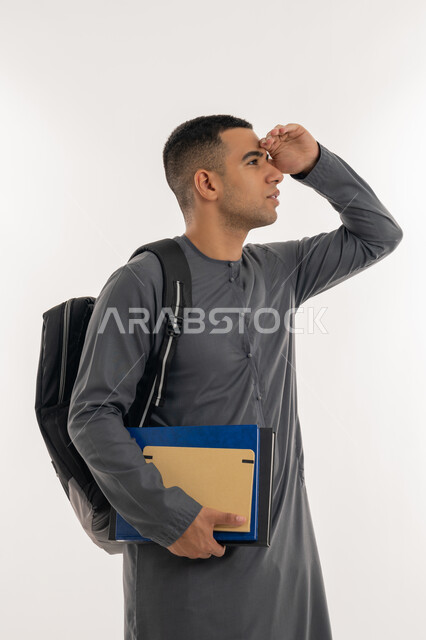 النظر لمكان بعيد, تعابير محاولة رؤية شيء ما, مفهوم التطلع للمستقبل, التعليم الجامعي في الإمارات, بورتريه لطالب جامعي عربي خليجي إماراتي يرتدي الكندورة يحمل حقيبة على ظهره والكتب الجامعية بيده, خلفية رمادية