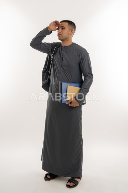 تعابير محاولة رؤية شيء ما, النظر لمكان بعيد, مفهوم التطلع للمستقبل, التعليم الجامعي في الإمارات, بورتريه لطالب جامعي عربي خليجي إماراتي يرتدي الكندورة يحمل حقيبة على ظهره والكتب الجامعية بيده, الجسم بالطول الكامل, خلفية رمادية
