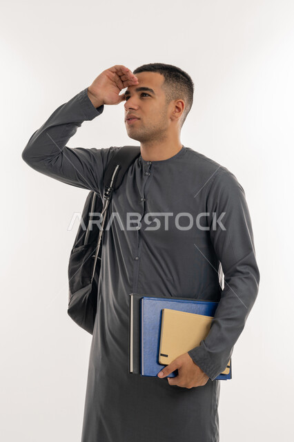النظر لمكان بعيد, تعابير محاولة رؤية شيء ما, مفهوم التطلع للمستقبل, التعليم الجامعي في الإمارات, بورتريه لطالب جامعي عربي خليجي إماراتي يرتدي الكندورة يحمل حقيبة على ظهره والكتب الجامعية بيده, خلفية رمادية