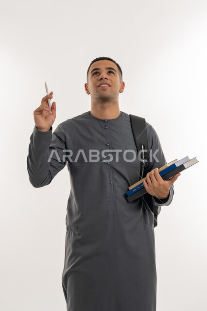 النظر إلى شيء ما بالأعلى, الاستعداد للعام الجامعي الجديد, التعليم العالي في الجامعات والكليات الأكاديمية الإماراتية, بورتريه لطالب جامعي عربي خليجي إماراتي يرتدي الكندورة يحمل حقيبة على ظهره وبيديه الكتب والقلم, خلفية رمادية