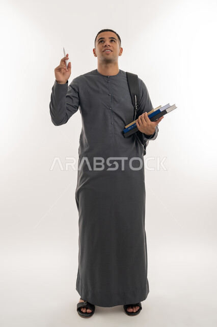 النظر إلى شيء ما بالأعلى, الاستعداد للعام الجامعي الجديد, التعليم العالي في الجامعات والكليات الأكاديمية الإماراتية, بورتريه لطالب جامعي عربي خليجي إماراتي يرتدي الكندورة يحمل حقيبة على ظهره وبيديه الكتب والقلم, الجسم بالطول الكامل, خلفية رمادية