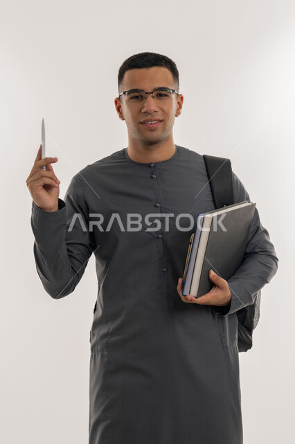 النظر إلى الكاميرا بإيماءات السعادة والسرور, الاستعداد للعام الجامعي الجديد, التعليم العالي في الجامعات والكليات الأكاديمية الإماراتية, بورتريه لطالب جامعي عربي خليجي إماراتي يرتدي الكندورة يحمل حقيبة على ظهره وبيديه الكتب والقلم, خلفية رمادية