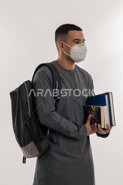 نشر الوعي الصحي, القيام بالإجراءات الوقائية لتجنب الإصابة بفايروس كورونا، التعليم الجامعي في الإمارات، بورتريه لطالب جامعي عربي خليجي إماراتي يرتدي الكندورة والكمامة يحمل حقيبة على ظهره وبيديه الكتب, خلفية رمادية