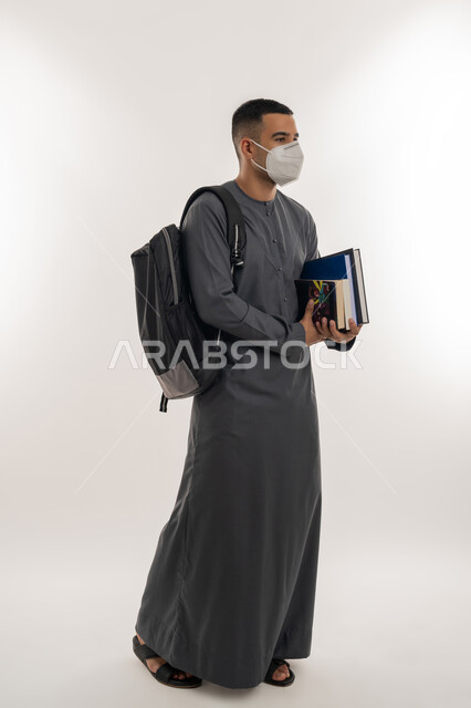 نشر الوعي الصحي, القيام بالإجراءات الوقائية لتجنب الإصابة بفايروس كورونا، التعليم الجامعي في الإمارات، بورتريه لطالب جامعي عربي خليجي إماراتي يرتدي الكندورة والكمامة يحمل حقيبة على ظهره وبيديه الكتب, الجسم بالطول الكامل, خلفية رمادية