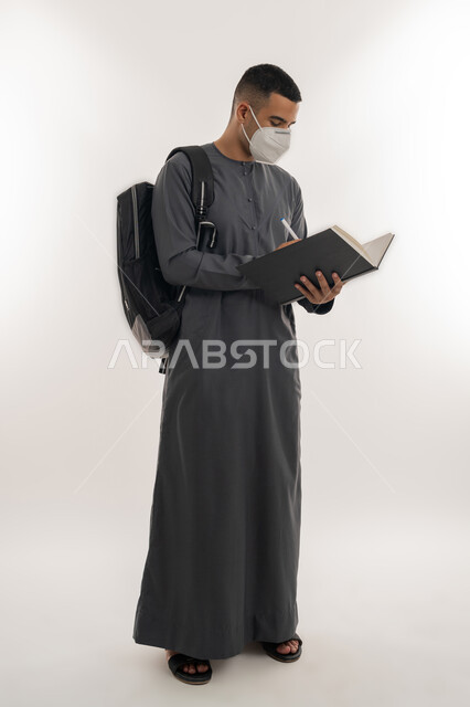 القيام بالإجراءات الوقائية لتجنب الإصابة بفايروس كورونا, نشر الوعي الصحي، التعليم الجامعي في الإمارات، بورتريه لطالب جامعي عربي خليجي إماراتي يرتدي الكندورة والكمامة يحمل حقيبة على ظهره ويقوم بتدوين الملاحظات, الجسم بالطول الكامل, خلفية رمادية