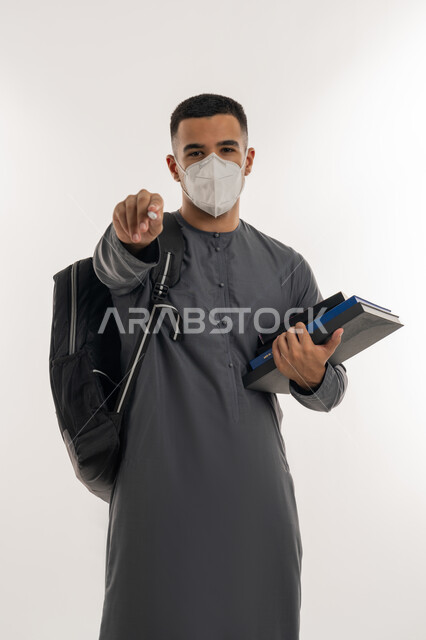 نشر الوعي الصحي للحد من انتشار الأمراض, اتباع الإجراءات الاحترازية للوقاية من فايروس كورونا، التعليم الجامعي في الإمارات، بورتريه لطالب جامعي عربي خليجي إماراتي يرتدي الكندورة والكمامة يحمل بيده الكتب ينظر إلى الكاميرا ويشير بإصبع السبابة للأمام, خلفية رمادية