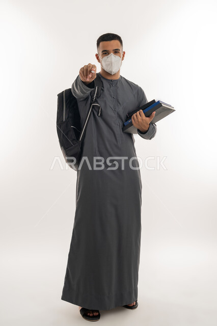 اتباع الإجراءات الاحترازية للوقاية من فايروس كورونا, نشر الوعي الصحي للحد من انتشار الأمراض، بورتريه لطالب جامعي عربي خليجي إماراتي يرتدي الكندورة والكمامة يحمل بيده الكتب ينظر إلى الكاميرا ويشير بإصبع السبابة للأمام, الجسم بالطول الكامل, خلفية رمادية