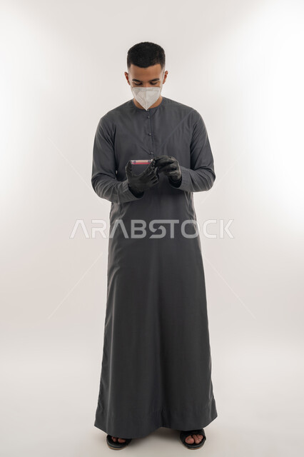 اتباع الإجراءات الاحترازية للوقاية من فايروس كورونا، نشر الوعي الصحي للحد من انتشار الأمراض، التطعيم الآمن لكوفيد19، بورتريه لطالب جامعي عربي خليجي إماراتي يرتدي الكندورة والكمامة يحمل بيده إبرة لقاح, الجسم بالطول الكامل، خلفية رمادية