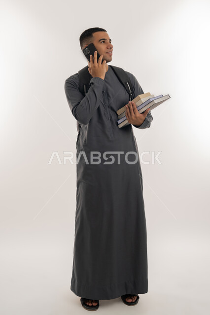 استخدام الأجهزة التقنية الحديثة, إجراء اتصال هاتفي عبر الجوال, التعليم الجامعي في الإمارات العربية المتحدة, بورتريه لطالب جامعي عربي خليجي إماراتي يرتدي الكندورة يحمل حقيبة على ظهره ويمسك بيديه الكتب والهاتف المحمول, الجسم بالطول الكامل, خلفية رمادية