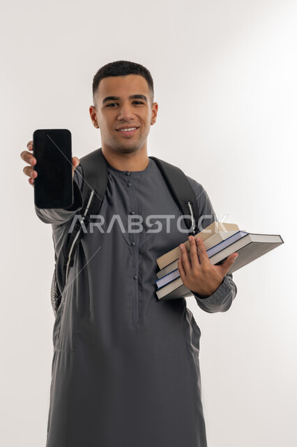 Looking at camera with gestures of happiness, using modern technology devices, university education in the Emirates, portrait of an Arab Gulf Emirati university student wearing a kandura carrying a bag on his back and holding books and a mobile phone in his hands, displaying a blank black screen via mobile, gray background