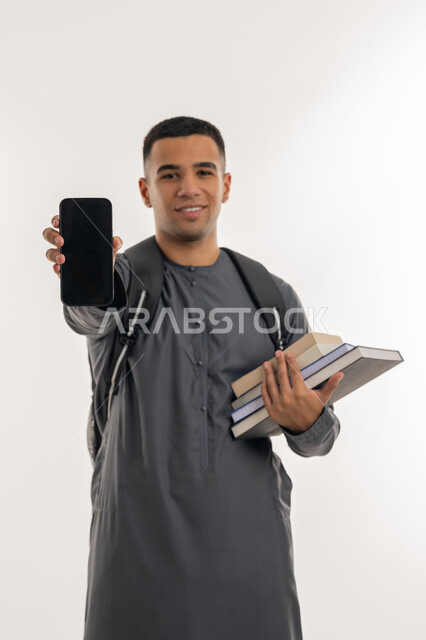 Looking at camera with gestures of happiness, using modern technology devices, university education in the Emirates, portrait of an Arab Gulf Emirati university student wearing a kandura carrying a bag on his back and holding books and a mobile phone in his hands, displaying a blank black screen via mobile, gray background