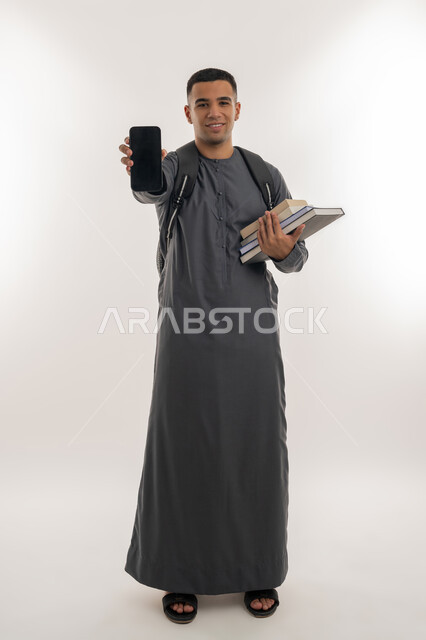 استخدام الأجهزة التقنية الحديثة, النظر إلى الكاميرا بإيماءات السعادة والسرور, بورتريه لطالب جامعي عربي خليجي إماراتي يرتدي الكندورة يحمل حقيبة على ظهره ويمسك بيديه الكتب والهاتف المحمول, عرض شاشة سوداء فارغة عبر الجوال, الجسم بالطول الكامل, خلفية رمادية
