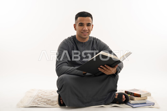 النظر إلى الكاميرا بإيماءات السعادة والسرور, التعليم الجامعي في الإمارات العربية المتحدة, مراجعة الدروس اليومية, بورتريه لطالب جامعي عربي خليجي إماراتي يرتدي الكندورة يجلس على الأرض يحمل بيده كتاب ويقوم بدراسة المحاضرات, خلفية بيضاء