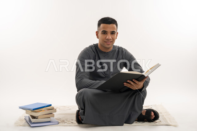 النظر إلى الكاميرا بإيماءات السعادة والسرور, التعليم الجامعي في الإمارات العربية المتحدة, مراجعة الدروس اليومية, بورتريه لطالب جامعي عربي خليجي إماراتي يرتدي الكندورة يجلس على الأرض يحمل بيده كتاب ويقوم بدراسة المحاضرات, خلفية بيضاء