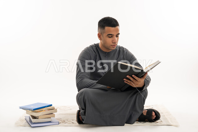 التعليم الجامعي في الإمارات العربية المتحدة, مفهوم الاجتهاد والتفوق, مراجعة الدروس اليومية, بورتريه لطالب جامعي عربي خليجي إماراتي يرتدي الكندورة يجلس على الأرض يحمل بيده كتاب ويقوم بدراسة المحاضرات, خلفية بيضاء