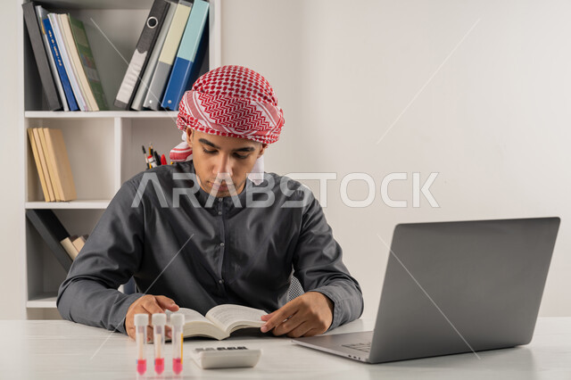دمج التقنيات الحديثة بالتعليم الأكاديمي, مفهوم الاجتهاد والتفوق, المناهج والمواد التعليمية, بورتريه مقرب لطالب جامعي عربي خليجي إماراتي يرتدي الكندورة والعمامة يجلس على الطاولة في مكتبة الجامعة ويقوم بمراجعة الدروس اليومية, خلفية رمادية