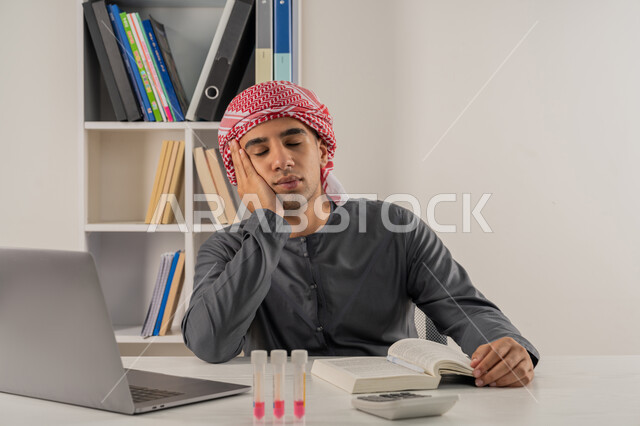 Concept of diligence and excellence, integration of modern technologies with academic education, curricula and educational materials, close-up portrait of an Arab Gulf Emirati university student wearing a kandura and turban sitting at a table in the university library and putting his hand on his cheek with gestures of fatigue and exhaustion, gray background