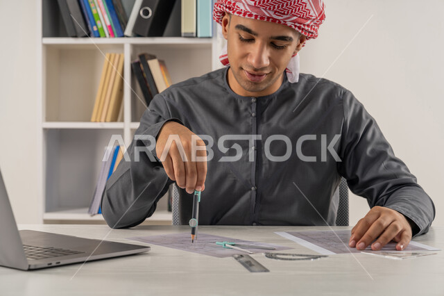 دمج التقنيات الحديثة بالتعليم الأكاديمي, مفهوم الاجتهاد والتفوق, المناهج والمواد التعليمية, بورتريه مقرب لطالب جامعي عربي خليجي إماراتي يرتدي الكندورة والعمامة يجلس على الطاولة في مكتبة الجامعة ويقوم بالرسم باستخدام الفرجار, خلفية رمادية