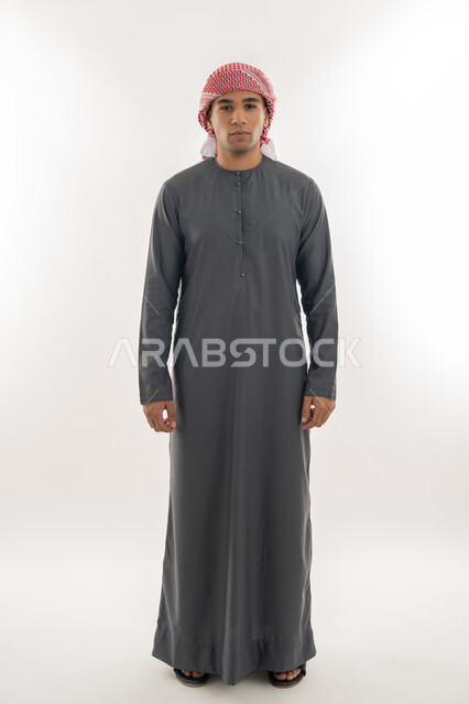 النظر إلى الكاميرا بإيماءات السعادة والسرور, الاهتمام بالمظهر الخارجي, مفهوم الأناقة والرجولة, بورتريه لشاب عربي خليجي إماراتي يرتدي الكندورة والعمامة يقف باستقامة واعتدال, الجسم بالطول الكامل, خلفية بيضاء