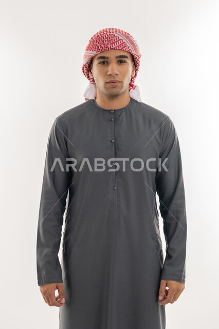النظر إلى الكاميرا بإيماءات السعادة والسرور, الاهتمام بالمظهر الخارجي, مفهوم الأناقة والرجولة, بورتريه لشاب عربي خليجي إماراتي يرتدي الكندورة والعمامة يقف باستقامة واعتدال, خلفية بيضاء