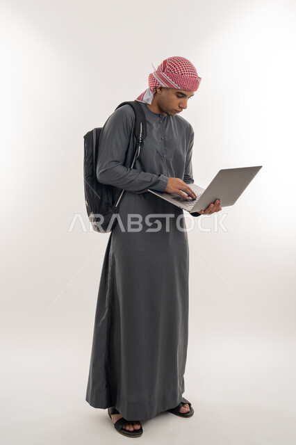 استخدام الأجهزة التقنية الحديثة لمتابعة المحاضرات الجامعية، مفهوم التعليم عن بعد، المناهج و المواد التعليمية في الإمارات, بورتريه لطالب جامعي عربي خليجي إماراتي يرتدي الكندورة و العمامة يحمل بيده الحاسوب المحمول, الجسم بالطول الكامل، خلفية بيضاء