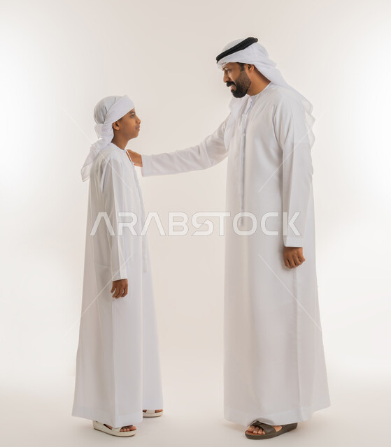 تعابير السعادة والسرور، تحمل المسؤولية والاعتناء بالأولاد، مفهوم الرابطة الأبوية والترابط الأسري، ايماءات الحب والعطف على الأبناء، بورتريه من الجانب لرجل عربي خليجي اماراتي يرتدي الكندورة والغترة يضع يده على كتف ابنه، صورة الجسم بالطول الكامل، خلفية بيضاء